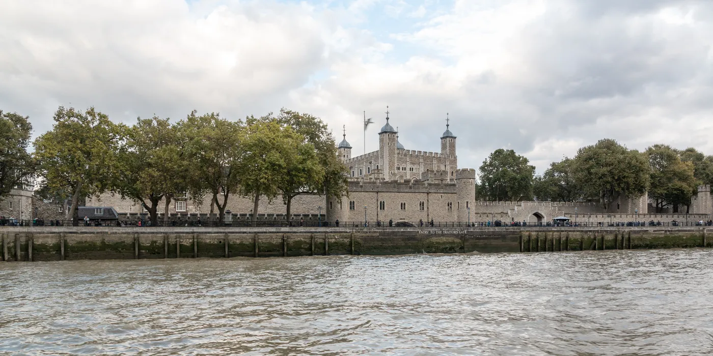 The Tower of London walls and White Tower seen across the River Thames, with trees along the wharf